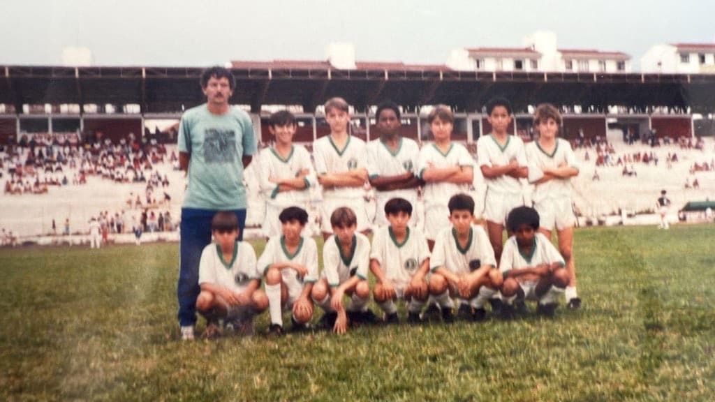 Youth team in white and green kits poses with their coach in front of a packed stadium grandstand