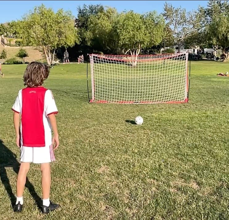 Young player in a red and white kit stands on the grass facing a portable goal with a ball at his feet